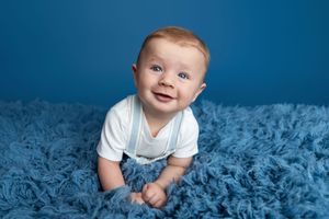 Smiling baby boy in white onesie with suspenders lying on blue fluffy blanket against blue background.