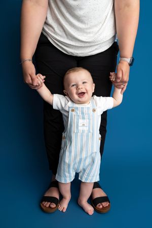 Happy baby in striped overalls stands supported by adult hands against a blue background.