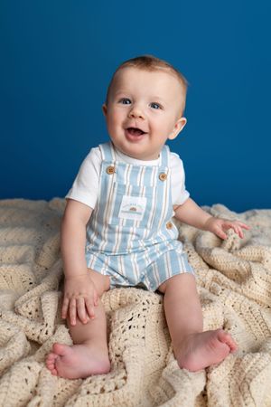 Smiling baby in striped overalls sits on cream crochet blanket against blue background.