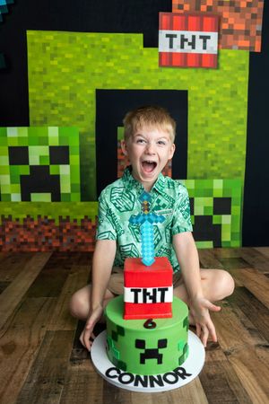 Excited boy celebrates 6th birthday with Minecraft-themed TNT cake and Creeper backdrop decorations.