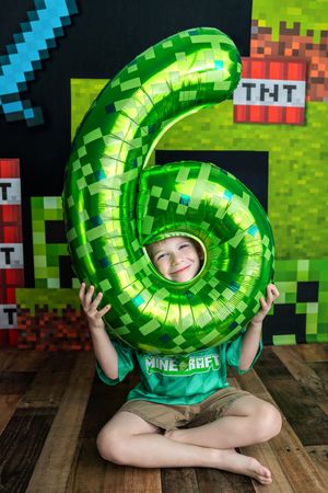 A smiling boy holds a green Minecraft-themed number 6 balloon at his birthday party with TNT blocks backdrop.