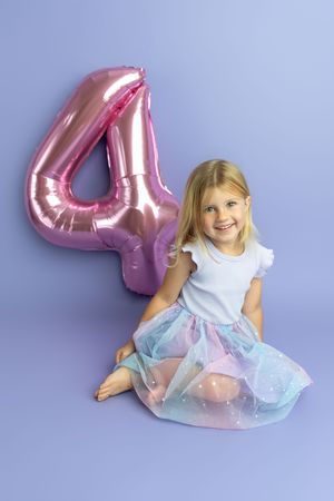 Smiling blonde girl in tutu skirt sitting beside pink number 4 balloon on purple background for birthday photo.