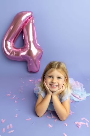 Smiling blonde girl lying on purple floor with pink number 4 balloon and confetti celebrating her 4th birthday.