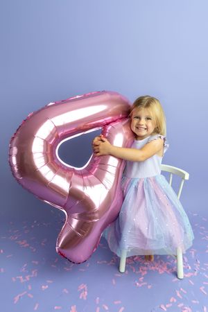 Smiling blonde girl in colorful dress hugging a pink number 4 balloon on purple background with confetti.