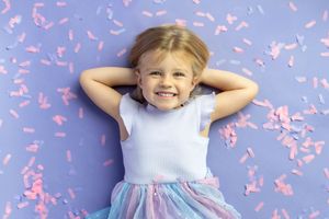 Happy little girl lying on purple background with pink confetti, smiling with hands behind her head.