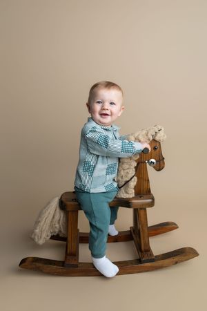 Smiling baby boy in teal outfit sitting on a wooden rocking horse toy against a beige background.