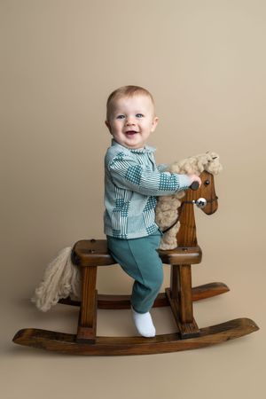 Smiling baby boy in teal outfit riding a wooden rocking horse toy against a beige background.