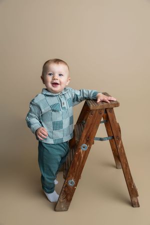 Smiling baby boy in teal patchwork shirt and pants standing beside a wooden step ladder on beige background.