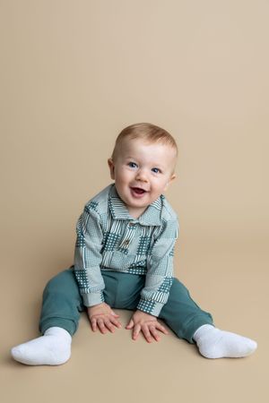 Happy baby boy sitting on beige background, wearing teal plaid shirt and green pants with white socks.