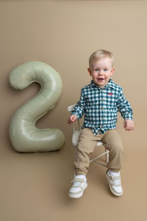 Smiling toddler boy in plaid shirt sits beside a sage green number 2 balloon on beige background.