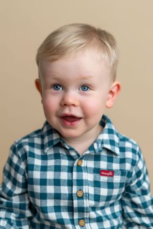 Smiling toddler boy with blue eyes and blonde hair wearing a teal plaid Wrangler button-up shirt.