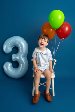 Happy toddler sitting on white chair celebrating 3rd birthday with colorful balloons and number 3 balloon on blue background.