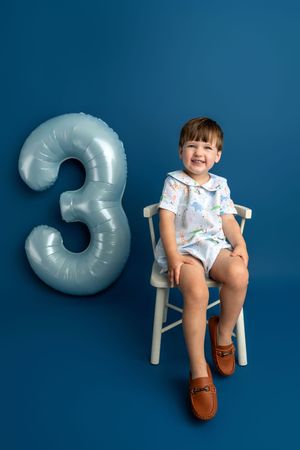 A smiling toddler boy sits on a white chair beside a silver number 3 balloon against a blue background.