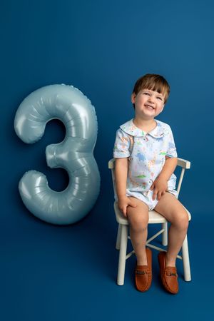 Happy toddler boy sitting on white chair next to number 3 balloon, celebrating 3rd birthday on blue background.