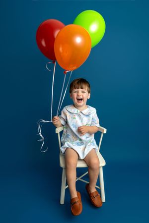 Laughing toddler sitting on white chair holding red, orange, and green balloons against blue background.