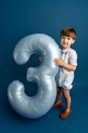 A toddler in a white outfit holds a large silver number 3 balloon against a blue background, celebrating their 3rd birthday.