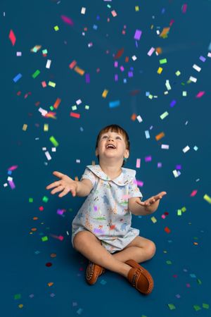 A joyful toddler girl in a floral dress sits cross-legged, laughing and reaching up as colorful confetti falls around her.