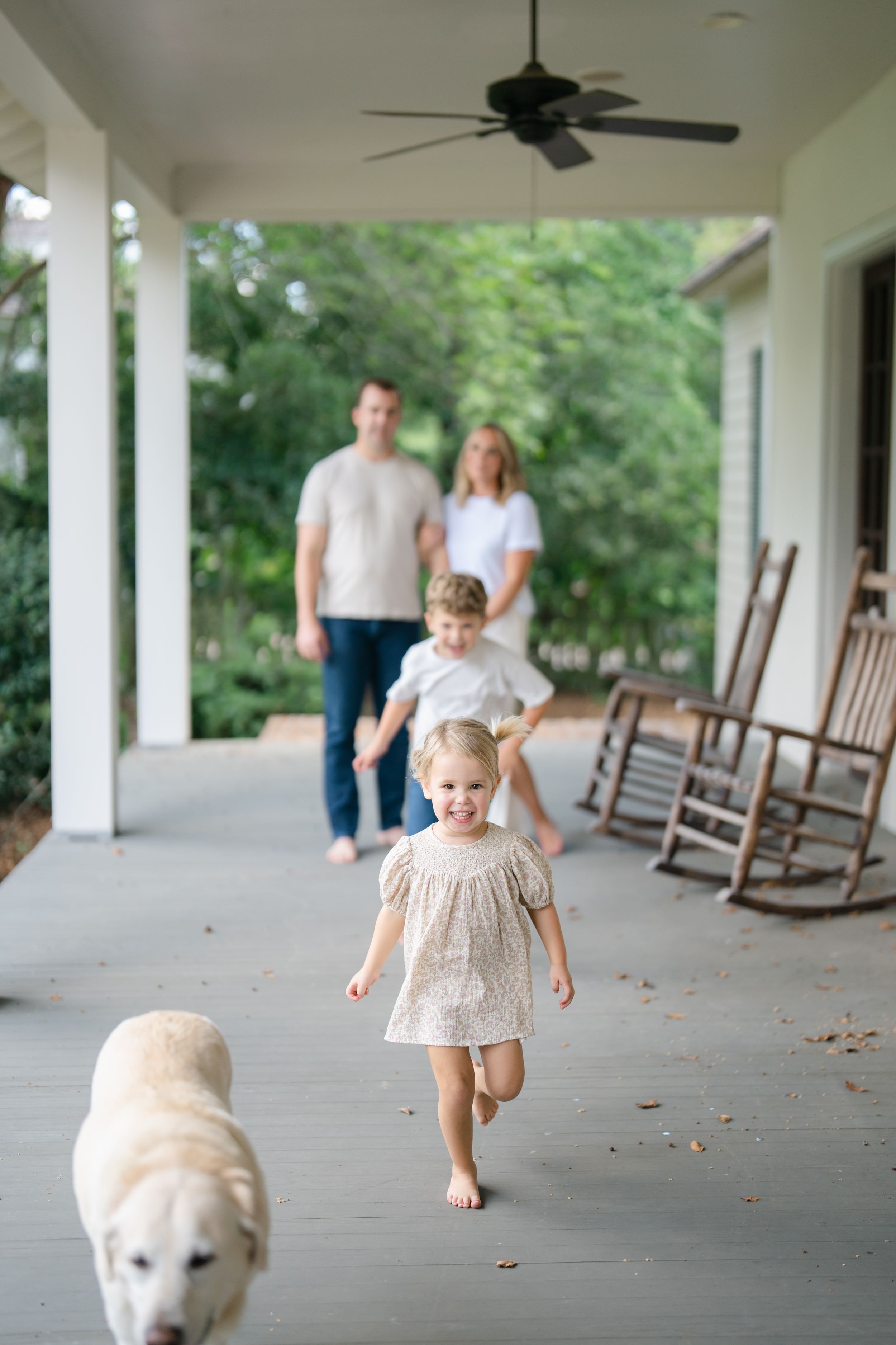 Young girl running barefoot on covered porch with family and golden dog in background.