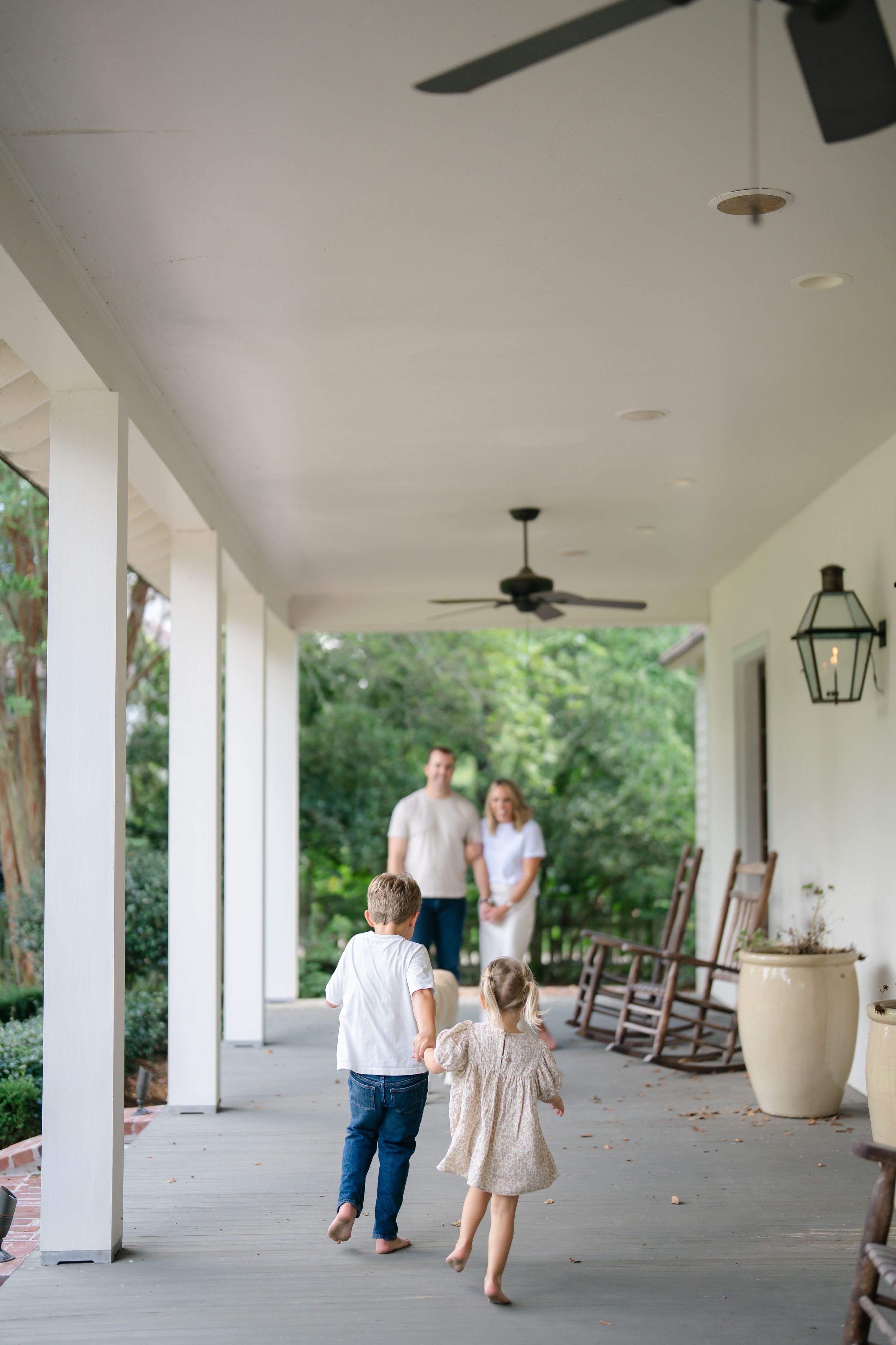 Two children holding hands walking toward parents on spacious white covered porch.