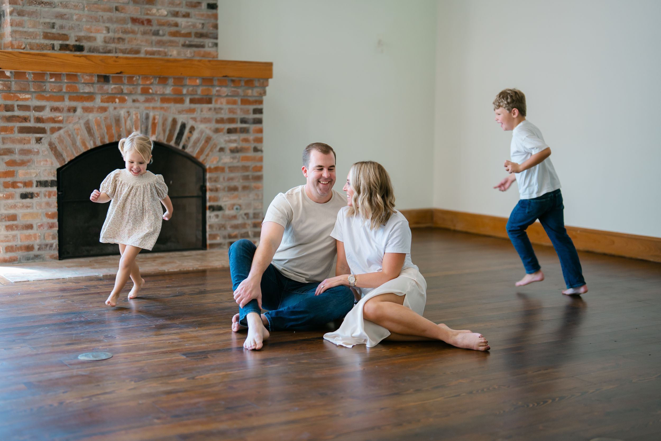 Happy family sitting on hardwood floor laughing while two young children play near brick fireplace.