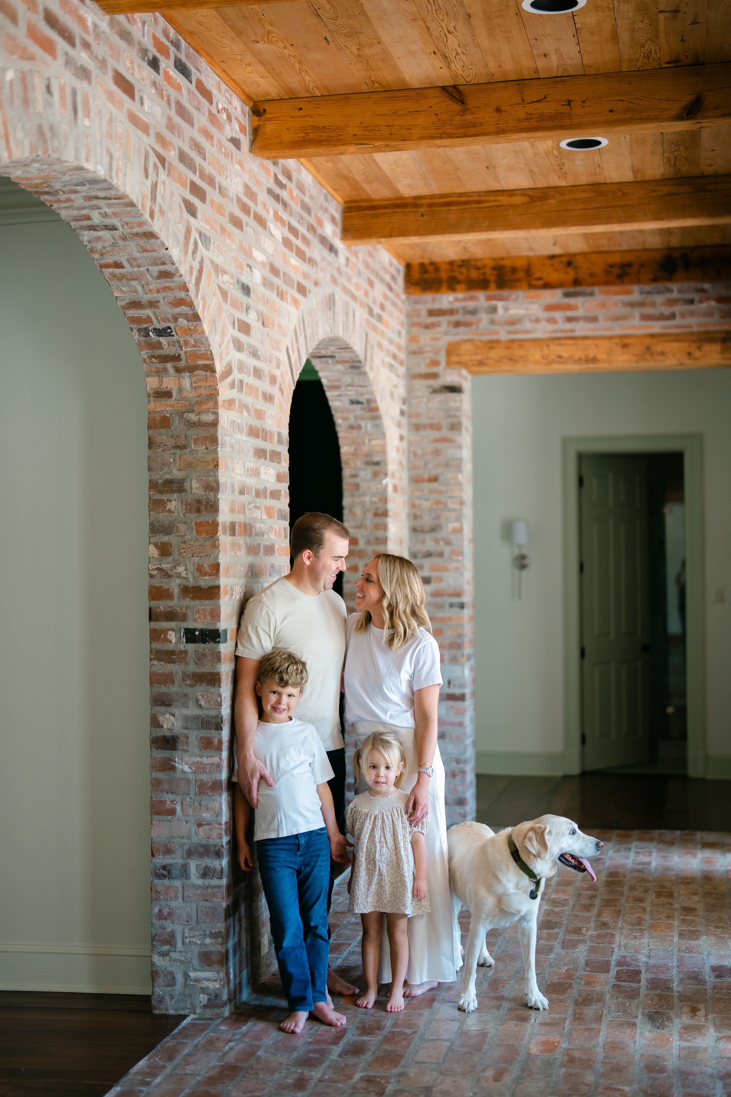 Family of four with dog posing under brick archway in rustic home with wood beam ceiling.