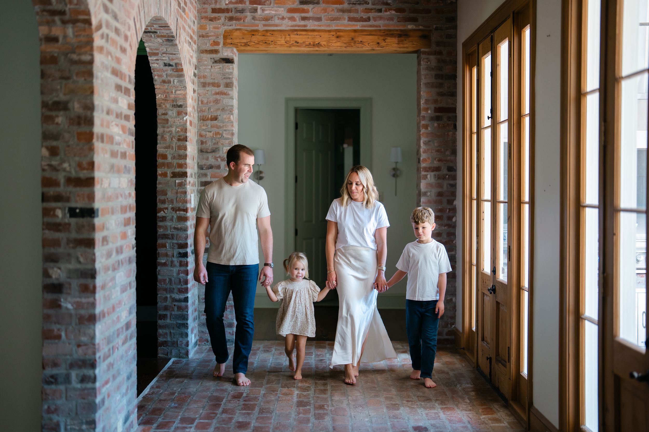 Family of four walking hand in hand through brick hallway of elegant home with arched doorways.