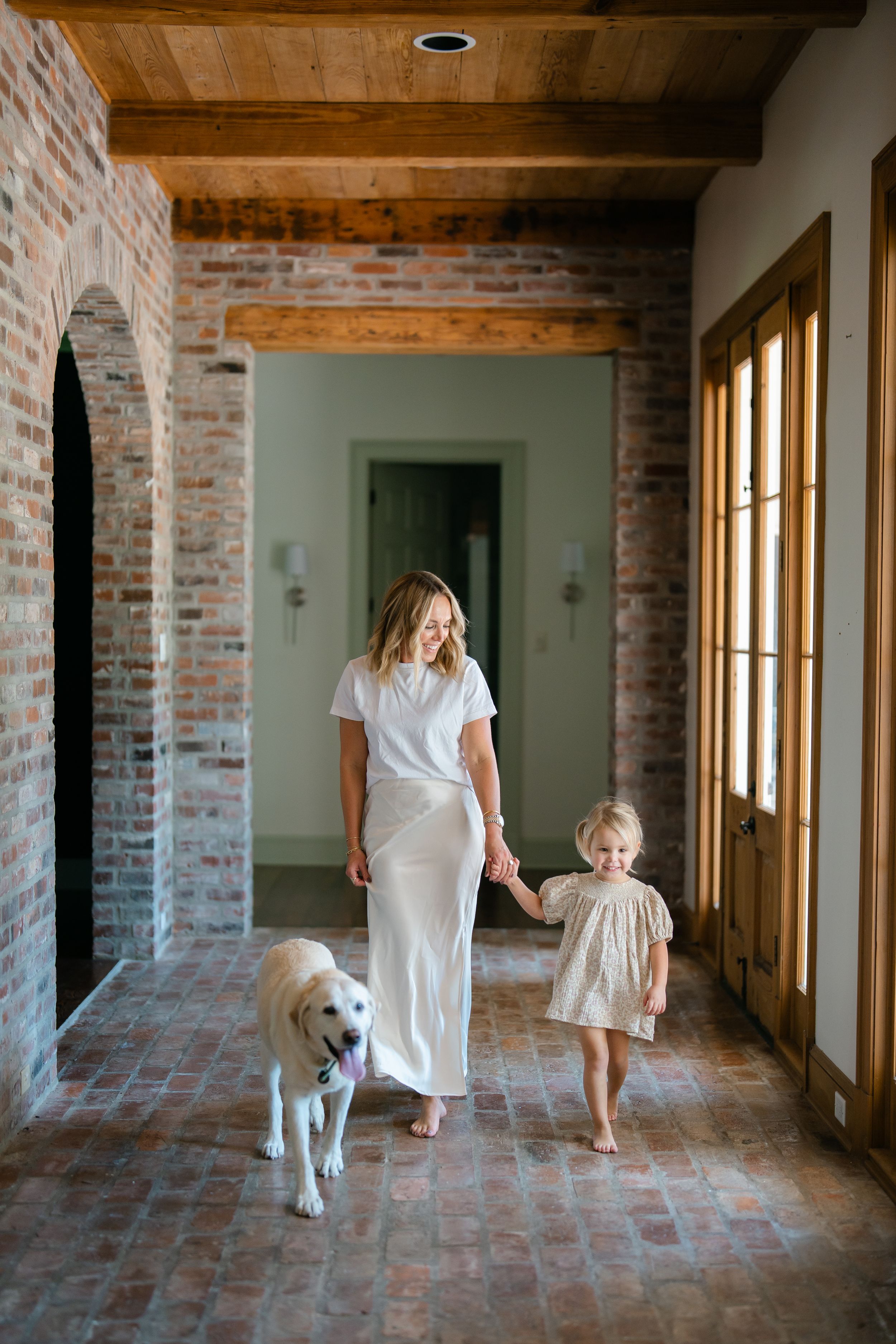 Mother in white skirt walking with young daughter and golden retriever through brick-floored home corridor.