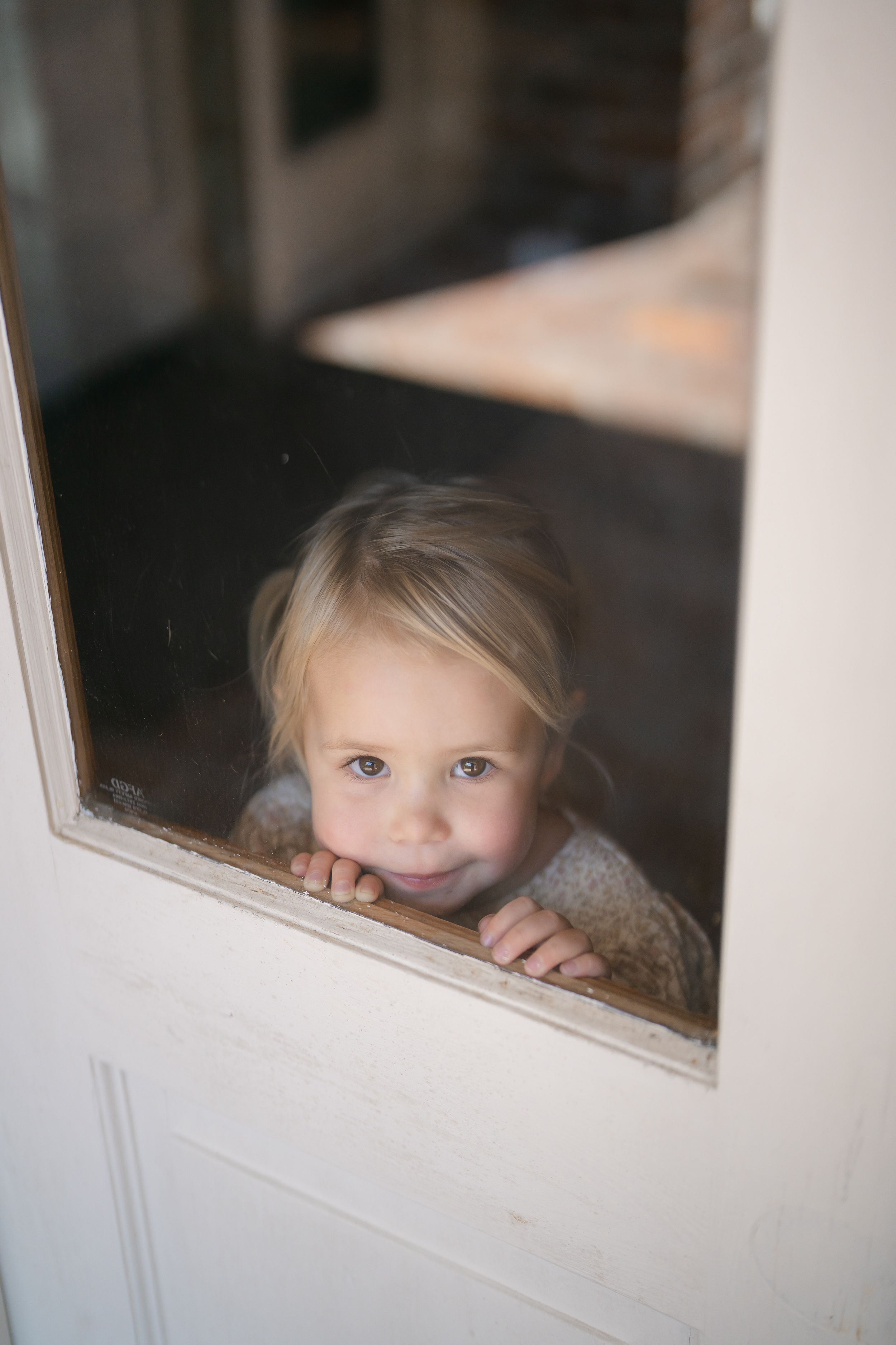 Adorable blonde toddler girl peering through glass window panel of white door with big smile.