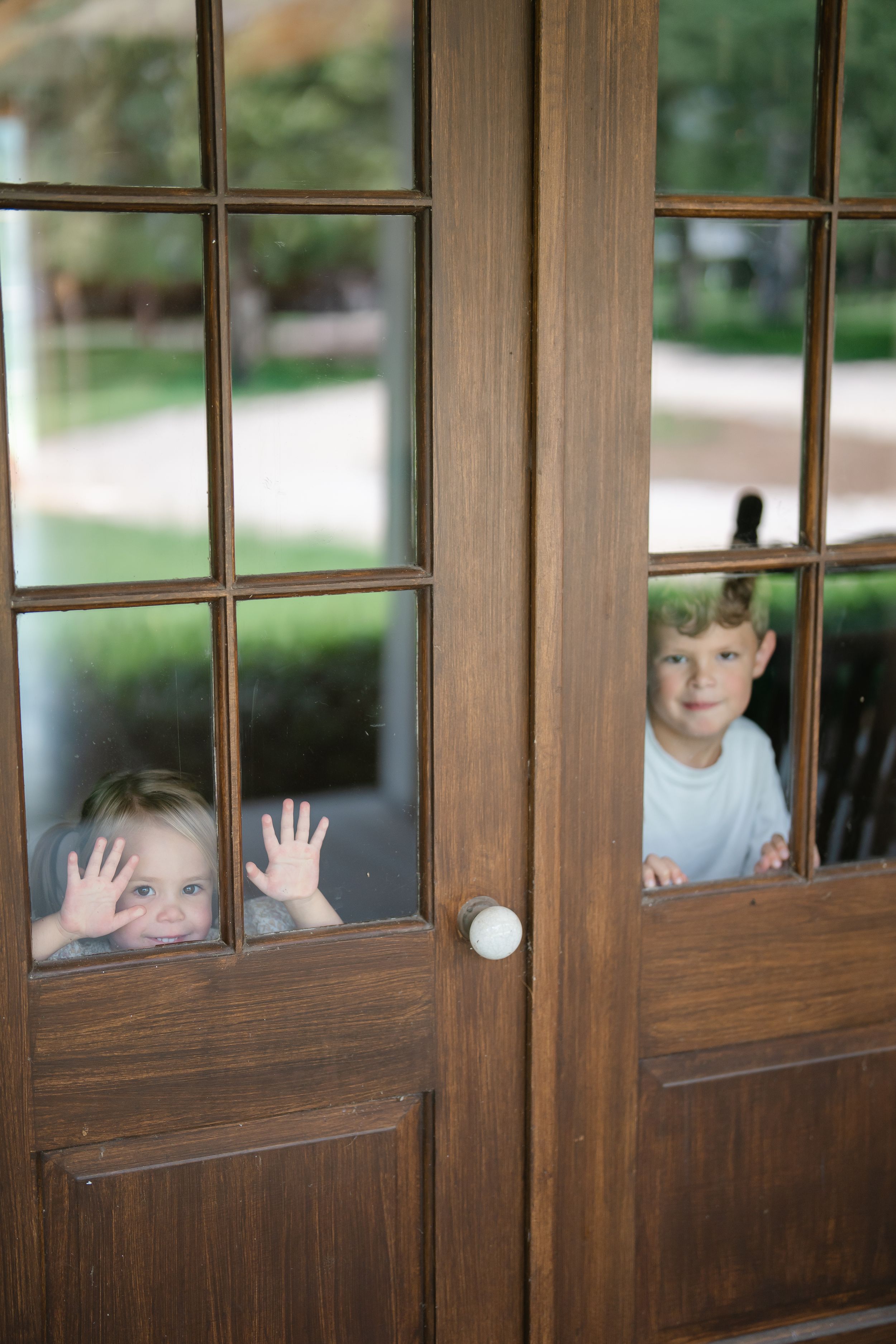 Two young children pressing faces and hands against glass French doors while looking inside playfully.