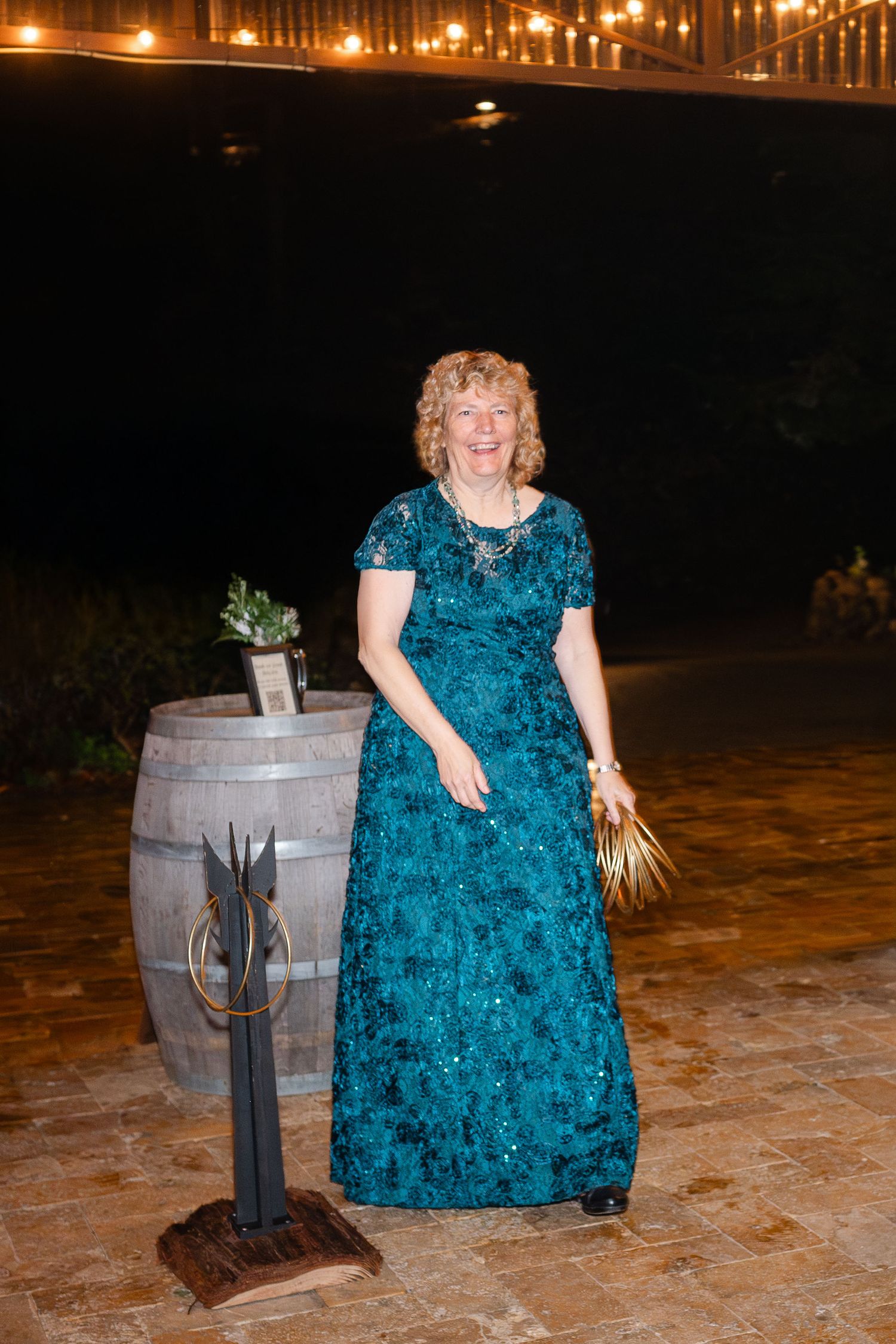 Woman in teal gown holds bouquet near rustic wine barrels at an outdoor evening wedding reception.