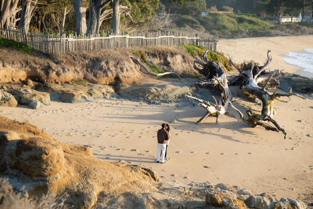 Couple embracing on a sandy beach near weathered driftwood, rocky cliffs and a wooden fence, with ocean waves in the distance.