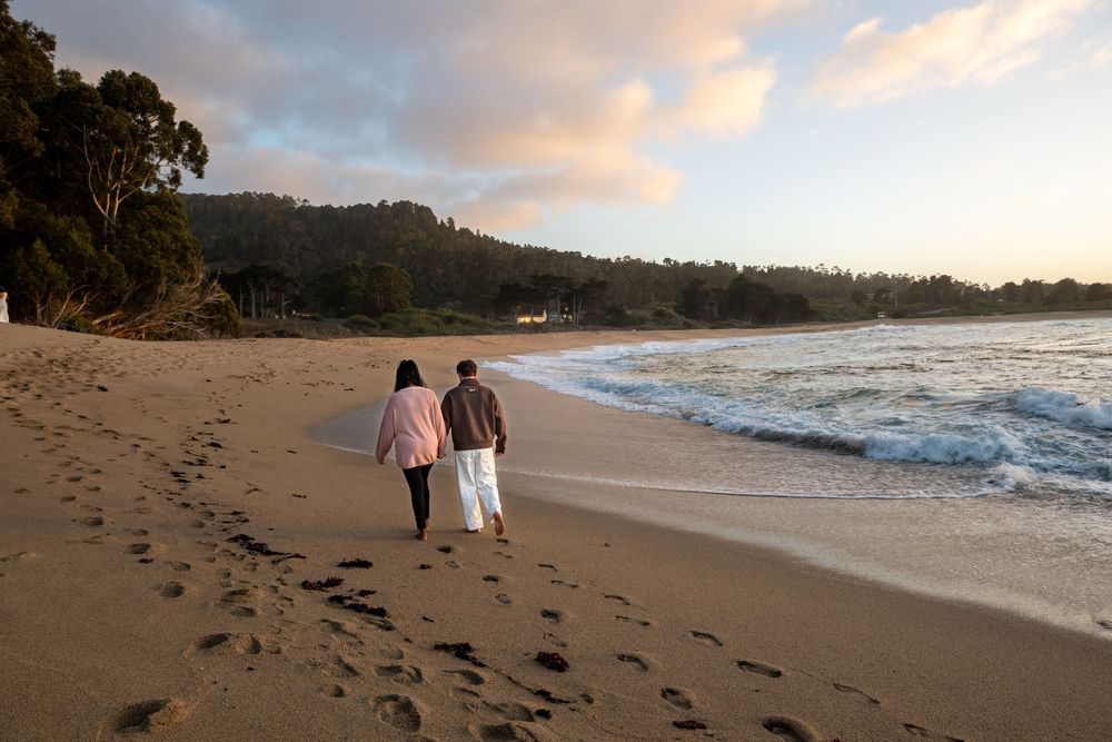 Two people walk together along a sandy beach at sunset, leaving footprints beside gentle ocean waves and a forested coastline.