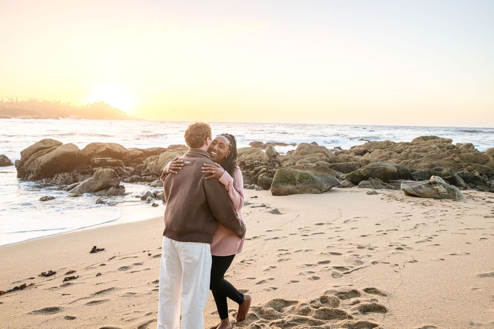 Couple embracing on a sandy beach at sunset with rocky shoreline and ocean waves in the background.