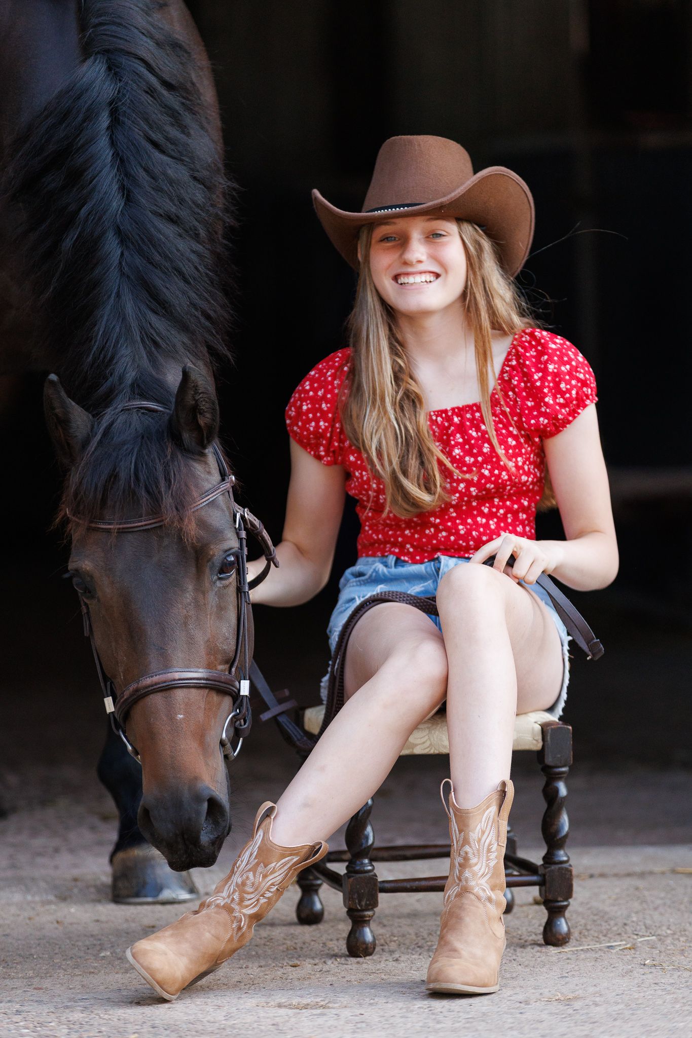 Smiling girl in a brown cowboy hat, red floral top and tan boots sits on a wooden stool beside a bridled brown horse in a barn.