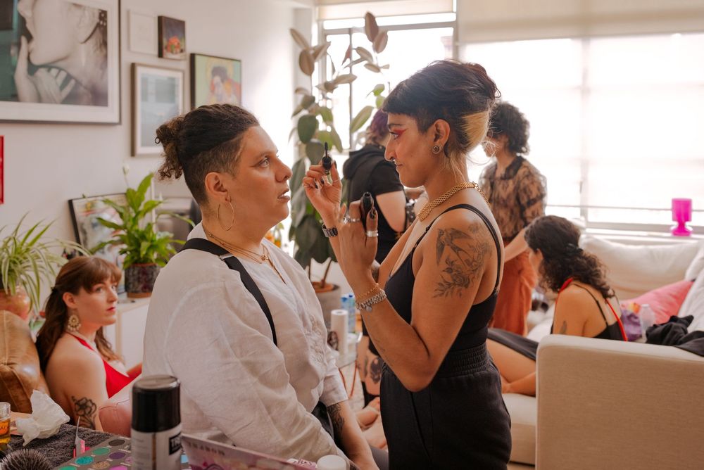 Two women interact in a lively indoor gathering, one applying makeup to the other, with tattoos and guests visible.
