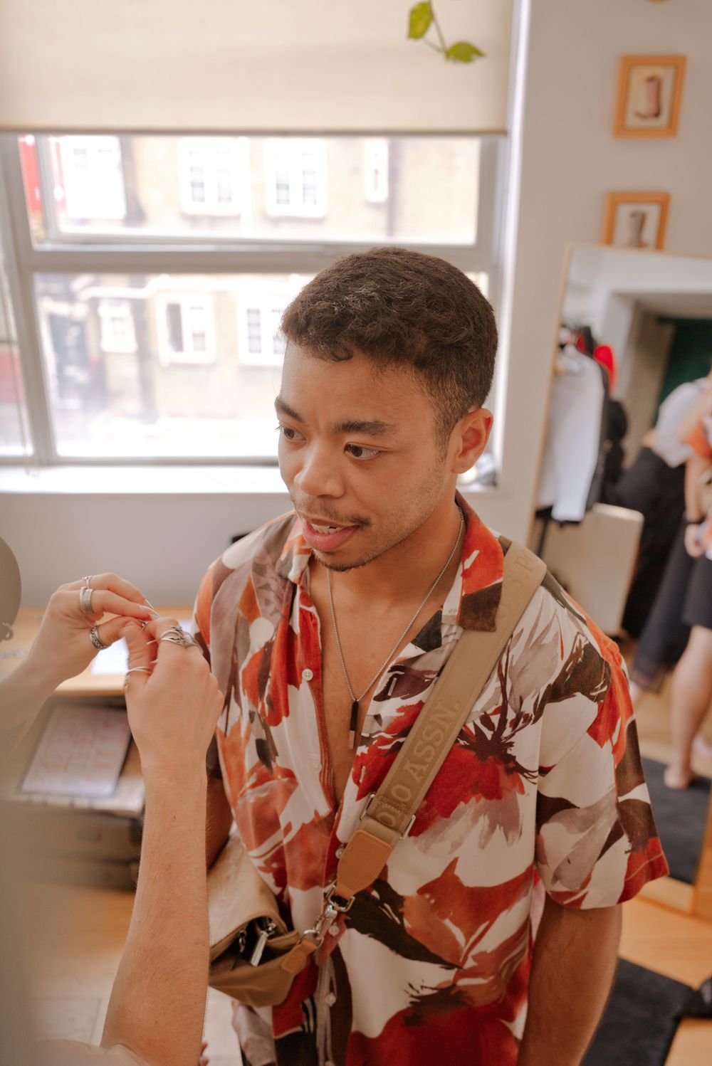 Young man in floral shirt smiling indoors, wearing a crossbody bag near a bright window.