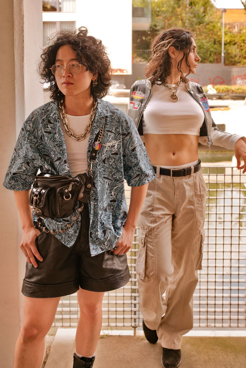 Two stylish women pose in urban setting, one in paisley shirt with belt bag, other in crop top and cargo pants.