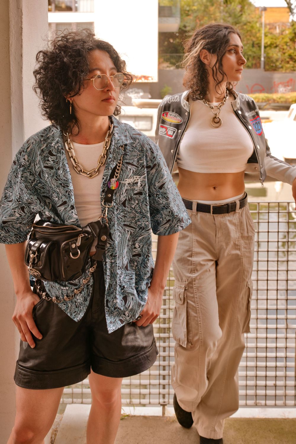 Two stylish women pose in urban setting wearing trendy outfits with chain necklaces, cargo pants, and patterned shirt.