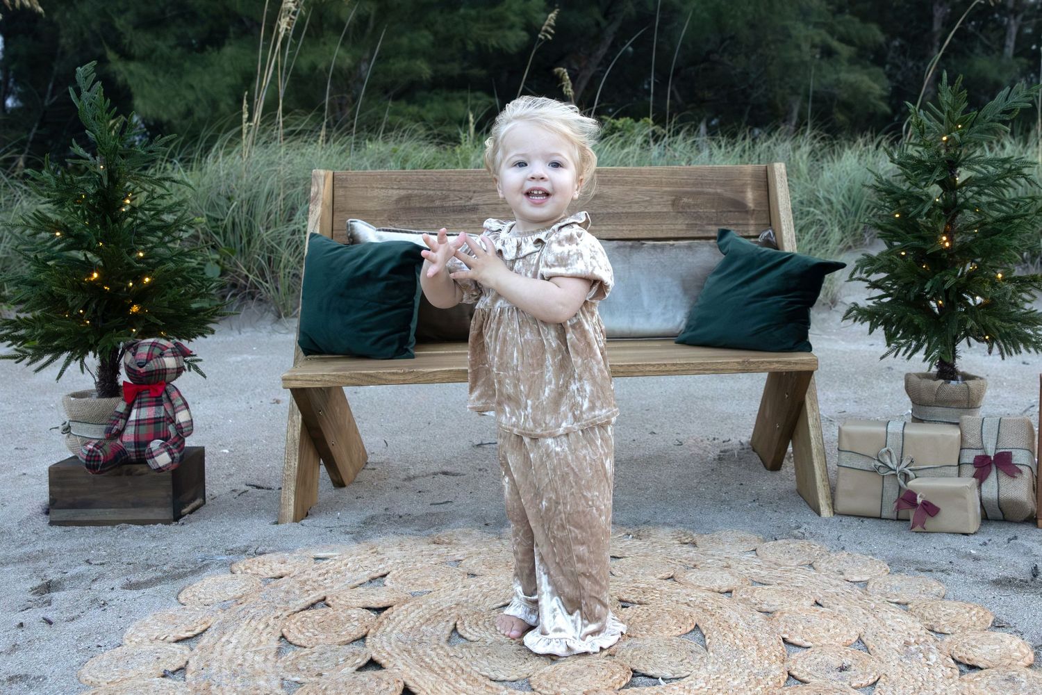 Toddler in a sparkly gold outfit stands on a woven mat at the beach beside a wooden bench, mini Christmas trees, and wrapped holiday gifts.