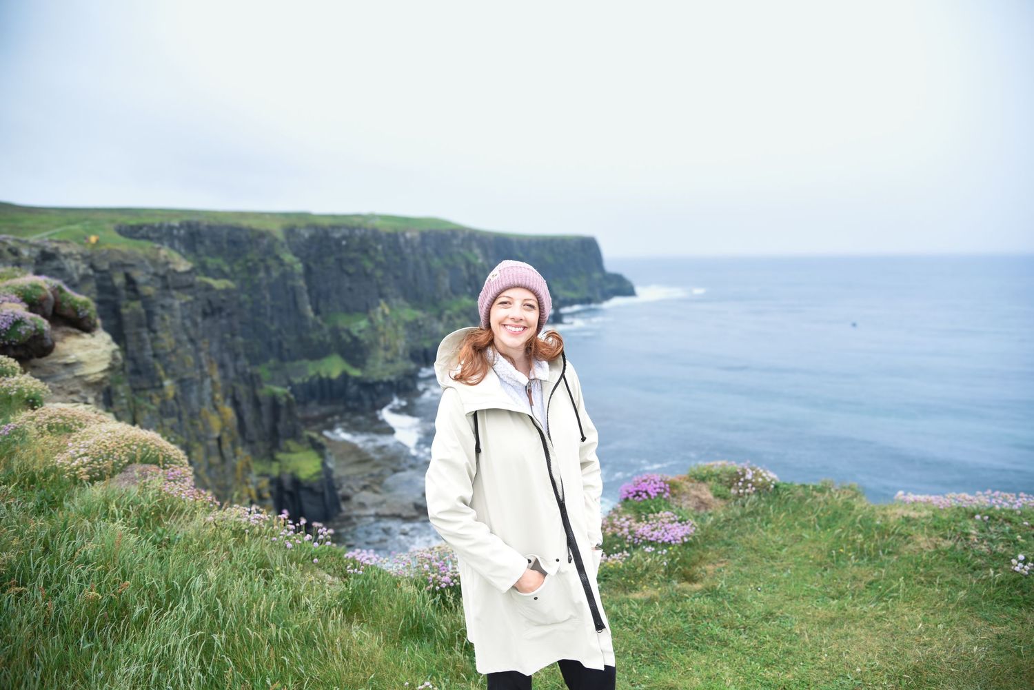 Smiling woman in a pink beanie and cream coat stands on a grassy clifftop with dramatic coastal cliffs and the ocean behind her.