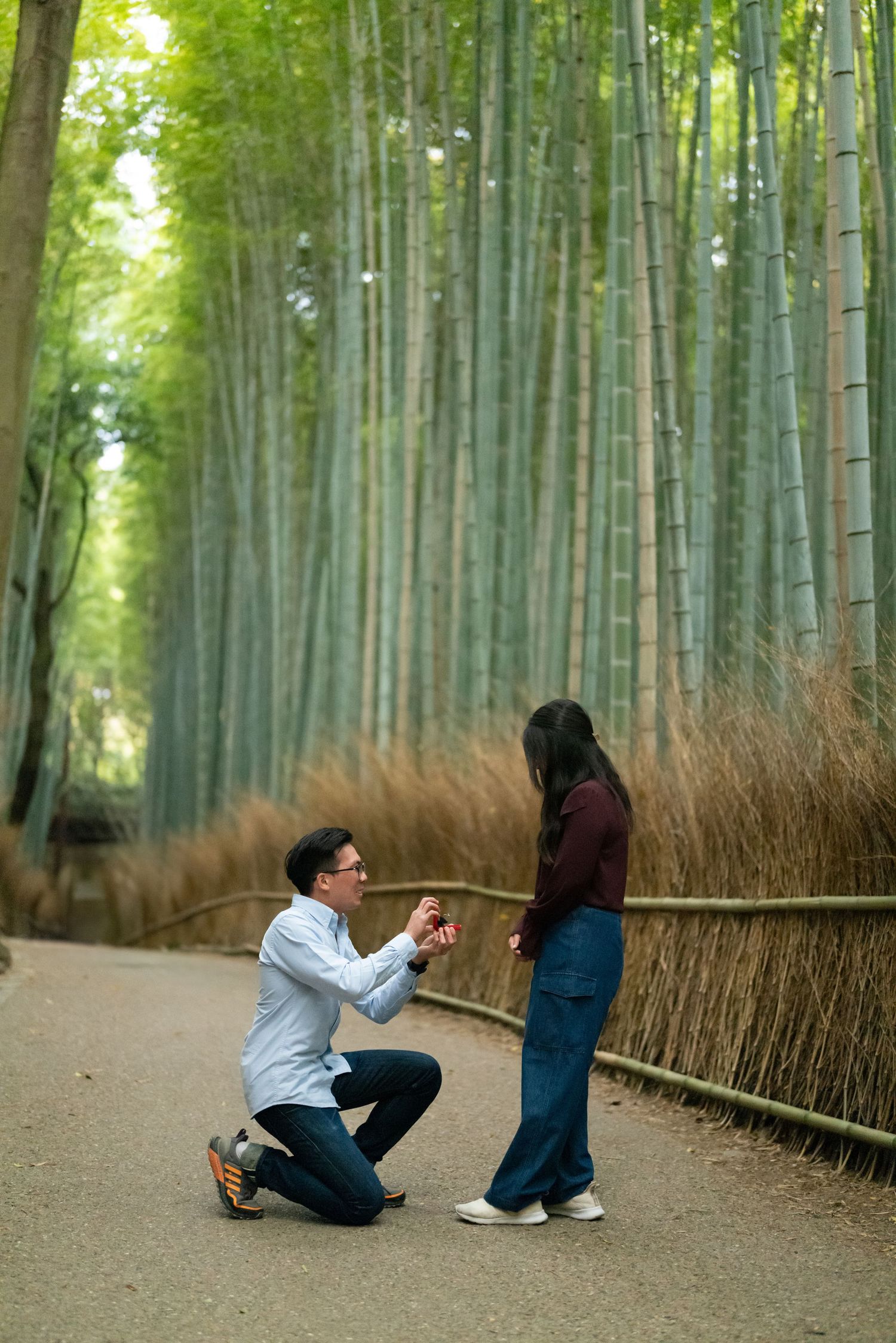 Man kneeling on one knee holds a ring while proposing to a woman on a path through a tall green bamboo forest in Kyoto.