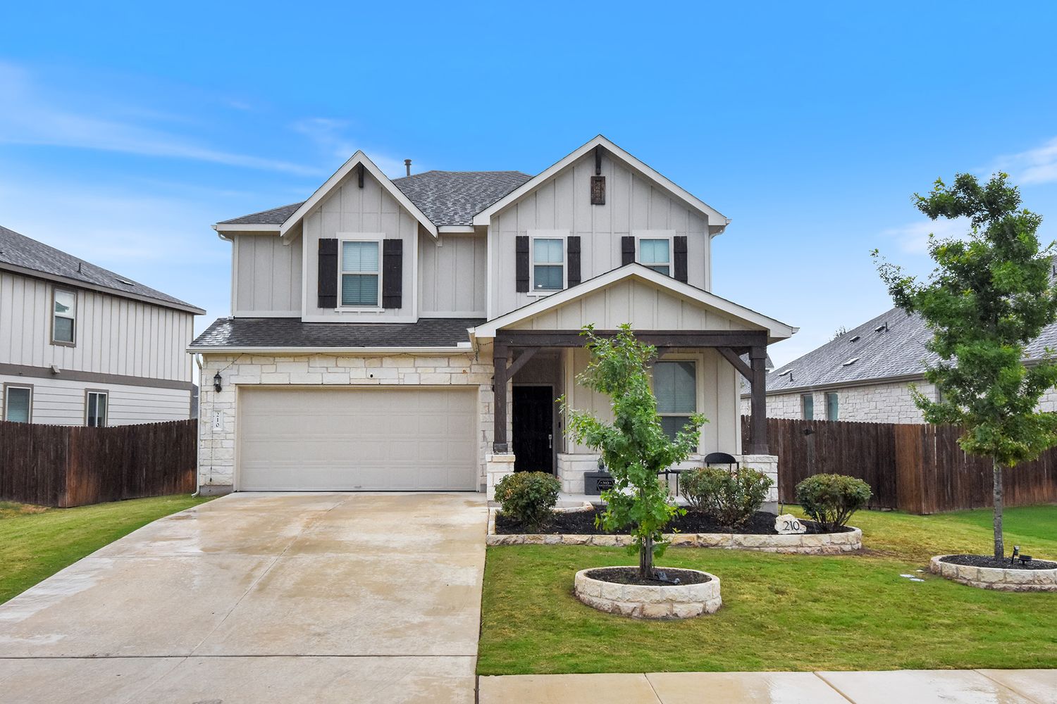 Two-story suburban home with tan siding, stone accents, covered front porch, two-car garage, and landscaped front yard with young tree.