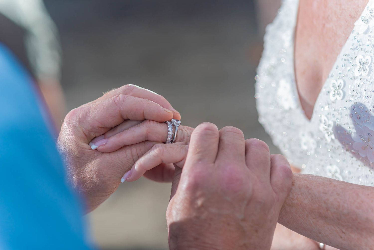 Close-up of hands exchanging a wedding ring during an outdoor ceremony, with a bride in a white lace dress standing nearby.