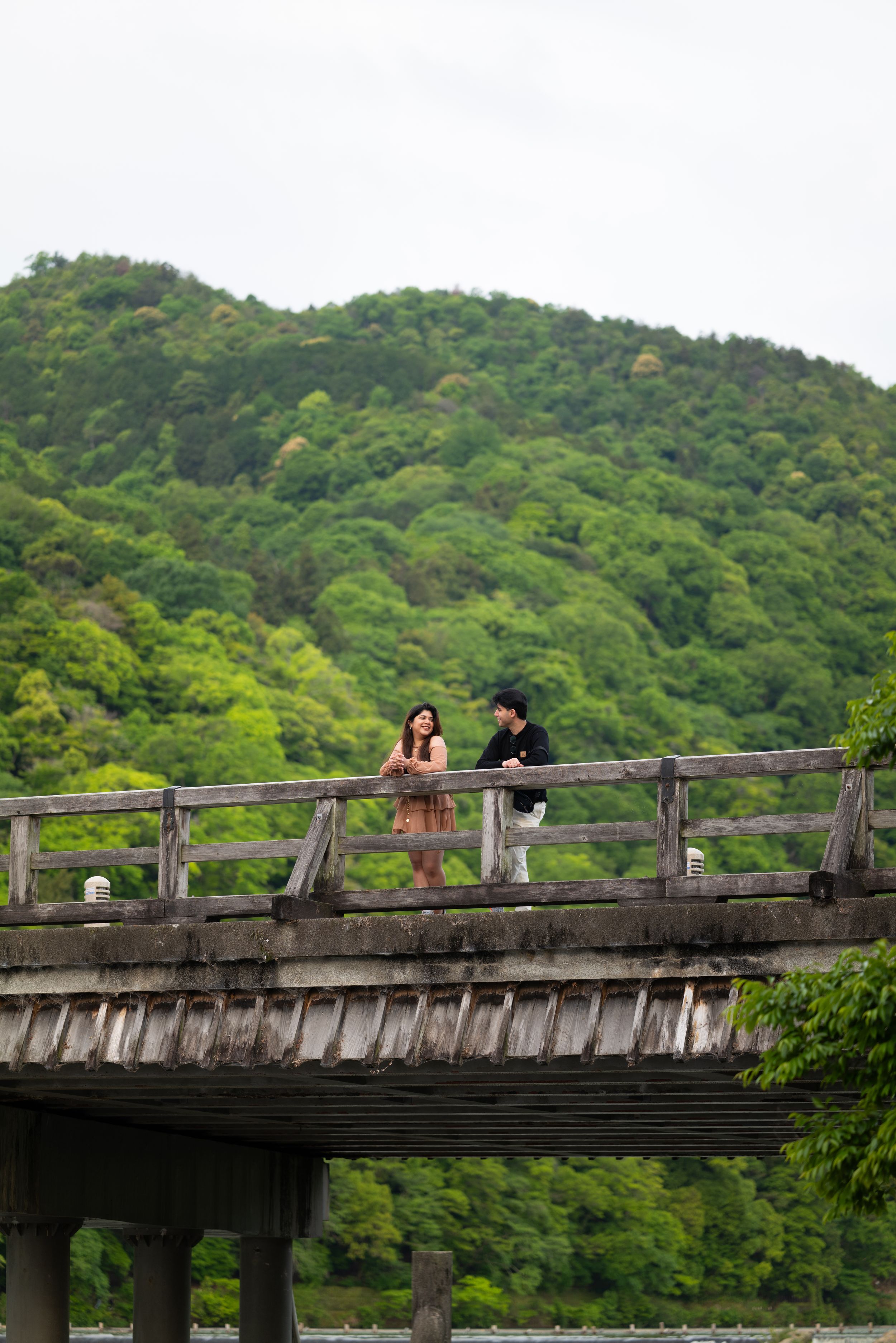 Two people stand side by side on a wooden bridge railing, looking out over a river with lush green forested mountains behind them.
