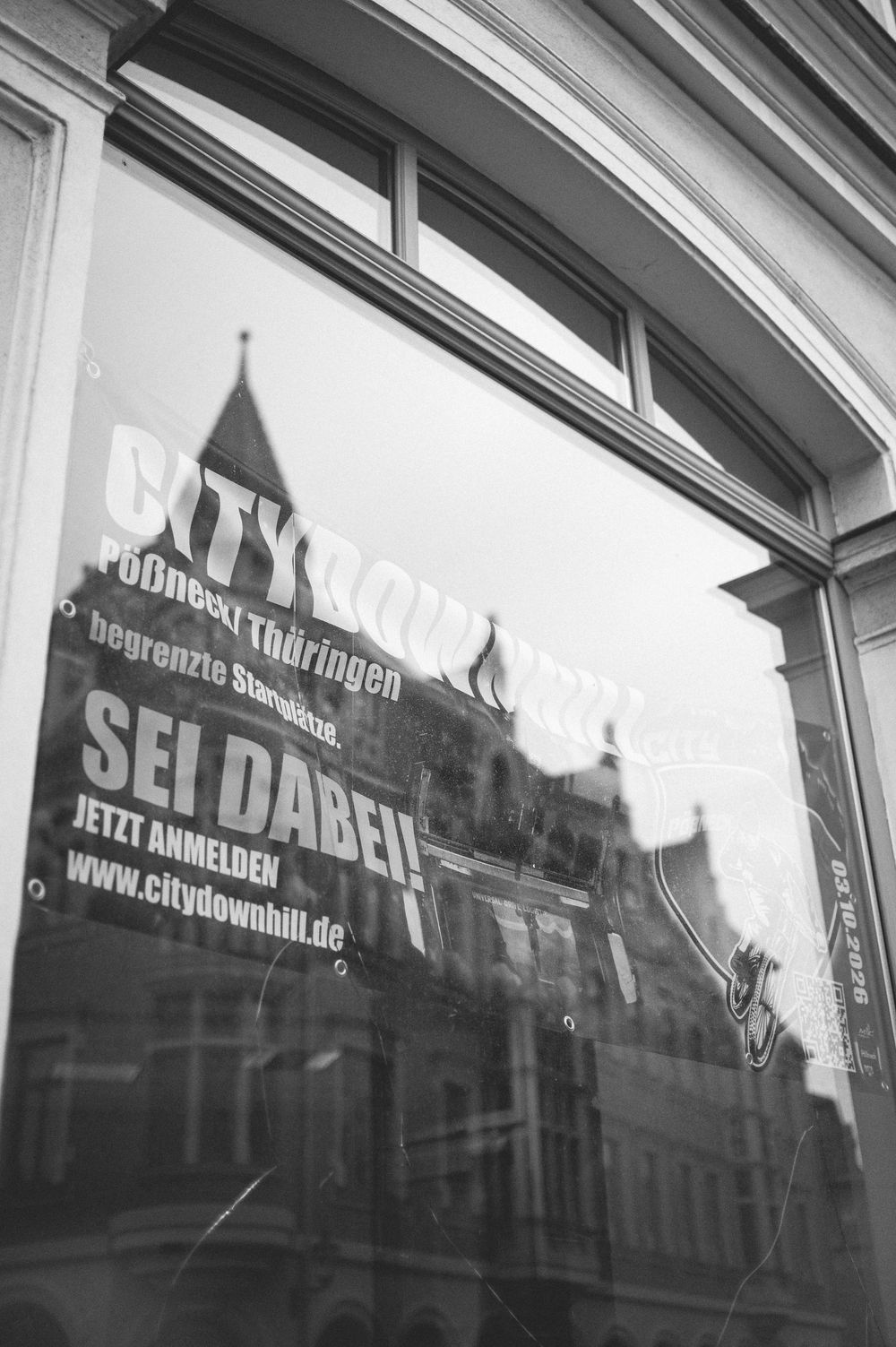 Black and white photo of a German City Downhill event poster on a window, reading SEI DABEI and www.citydownhill.de.