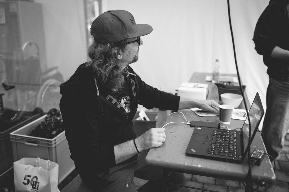 Black and white photo of a person with long hair wearing a cap, working on a laptop at a desk indoors.
