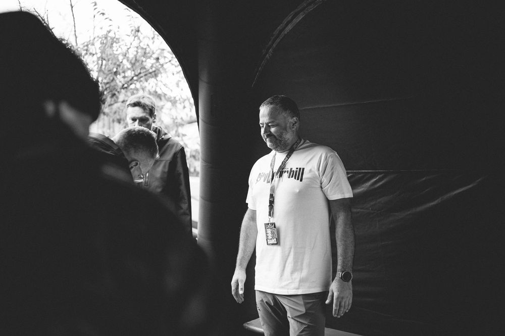 Black and white photo of two men standing under an archway outdoors, one wearing a white t-shirt with a lanyard.