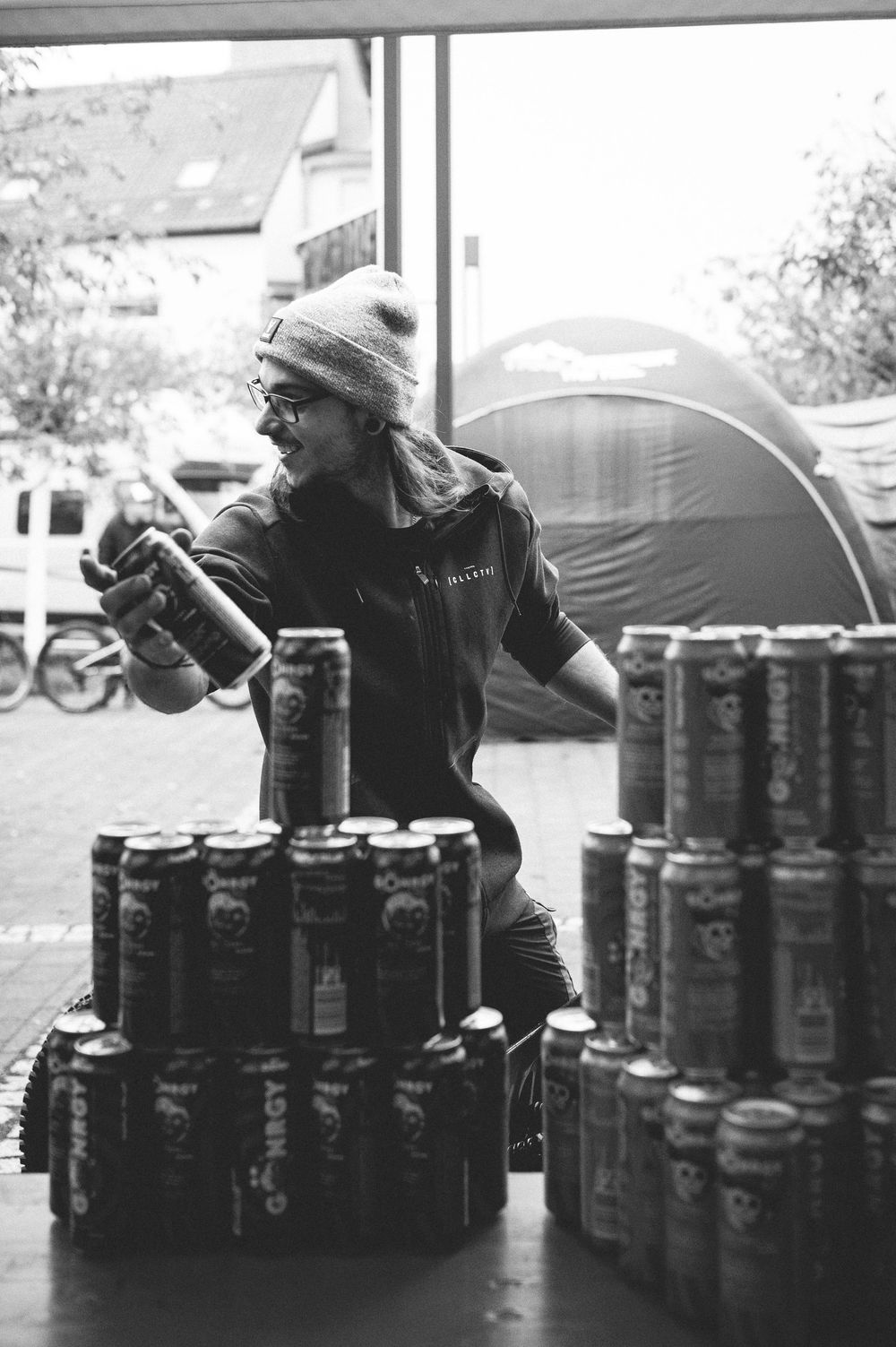 Man in beanie arranging stacked energy drink cans near a window in black and white photo.