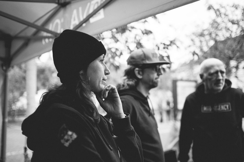 Black and white photo of three people standing under a tent outdoors, woman in foreground wearing black hoodie and beanie.