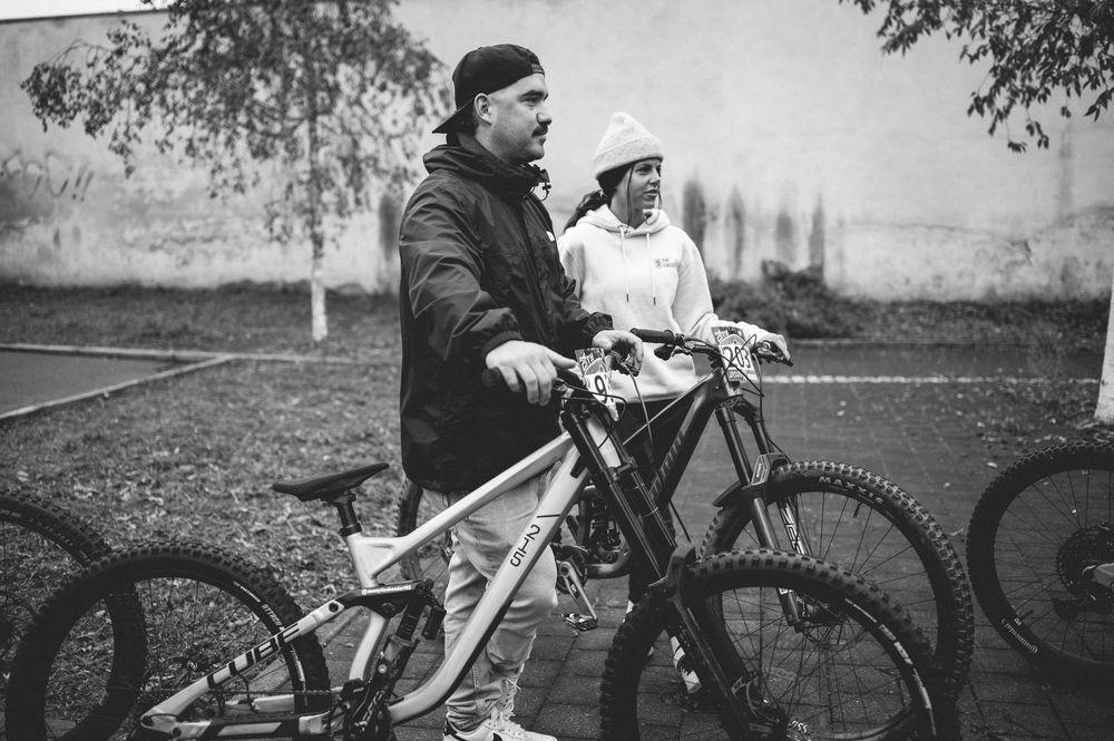 Two cyclists stand with mountain bikes outdoors in a black and white photo, wearing casual streetwear.